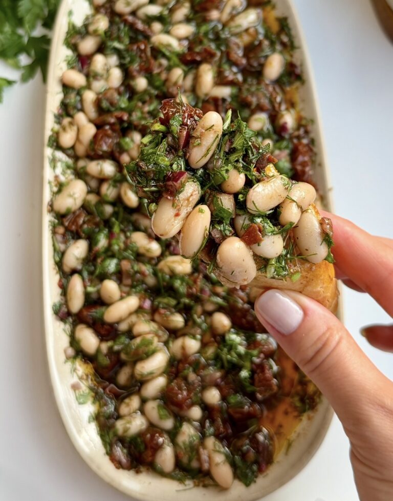 A hand holding toasted bread topped with olive oil white bean dip, above an oval platter of the dip made with cannellini beans, sun-dried tomatoes, red onion, parsley and dill. Lemons and fresh parsley sit in the background.