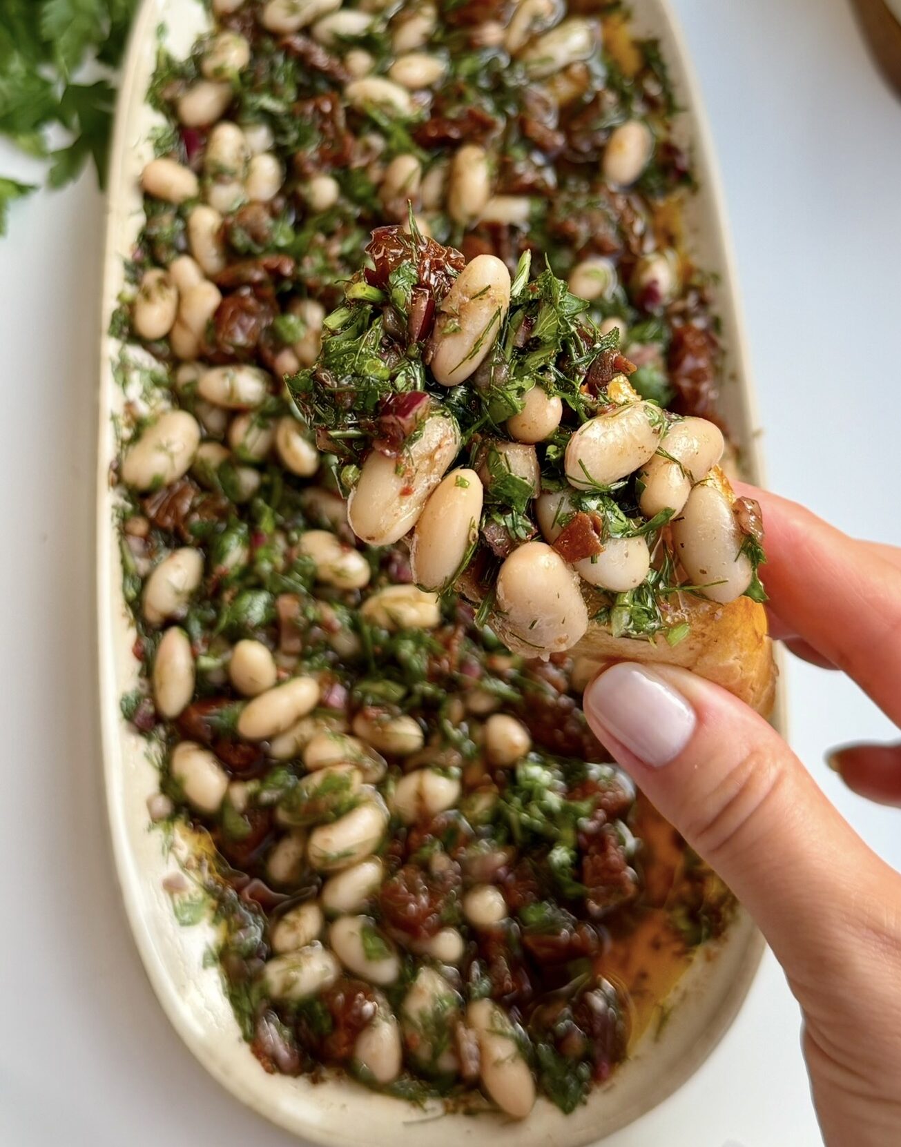 A hand holding toasted bread topped with olive oil white bean dip, above an oval platter of the dip made with cannellini beans, sun-dried tomatoes, red onion, parsley and dill. Lemons and fresh parsley sit in the background.