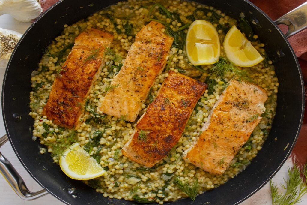 High protein meal prep idea: one pan salmon and pearl couscous with spinach, fresh dill, and lemon wedges