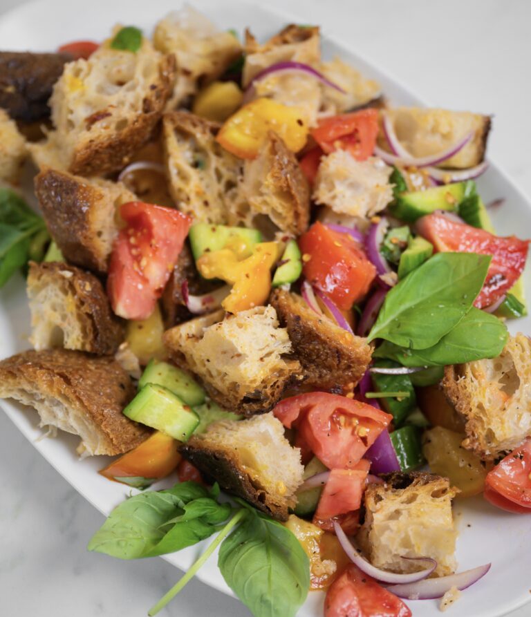 Panzanella salad with toasted bread cubes, red and yellow tomatoes, cucumber, red onion and fresh basil on a white platter.