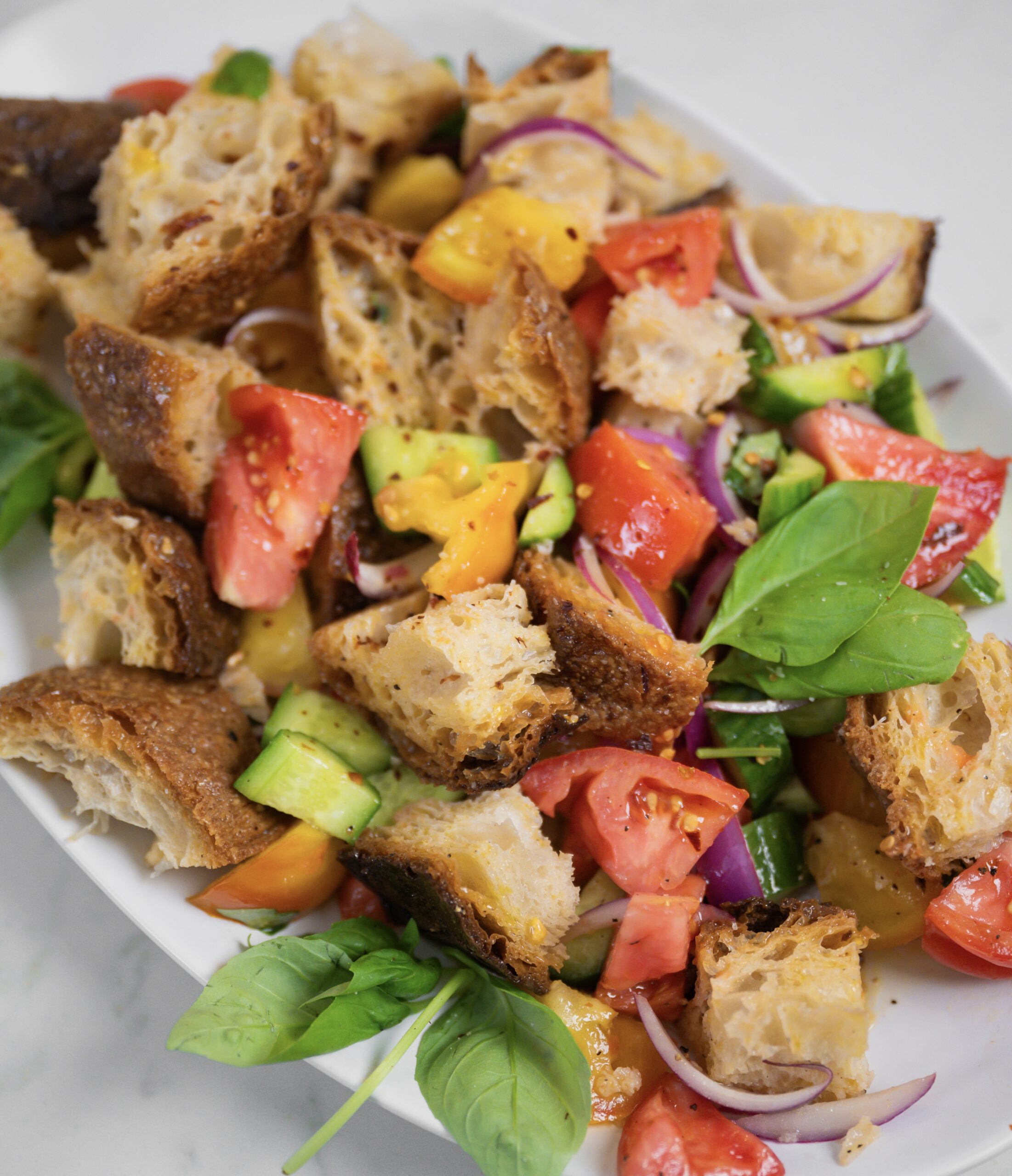 Panzanella salad with toasted bread cubes, red and yellow tomatoes, cucumber, red onion and fresh basil on a white platter.