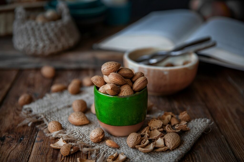 Green ceramic cup filled with in-shell almonds on burlap, with cracked shells scattered on a rustic wooden table, a nutrient-dense snack studied as almonds for sleep