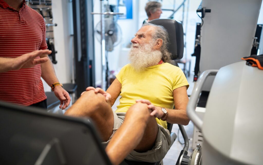 Older man on a leg press machine talking with a trainer, training to fight age-related muscle loss