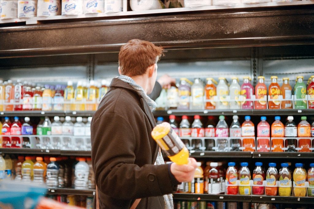 Shopper holding a bottled drink in front of a store cooler of sodas and juices, looking for the best protein soda