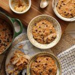 Bowls of morning glory oats with grated carrots and currants, shown with brown sugar, coconut flakes, and a pot of carrot cake oatmeal