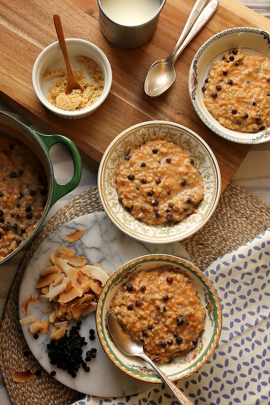 Bowls of morning glory oats with grated carrots and currants, shown with brown sugar, coconut flakes, and a pot of carrot cake oatmeal