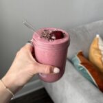 Hand holding a glass jar of cherry tahini smoothie topped with cacao nibs, sesame seeds, and a whole dark cherry, with a glass straw