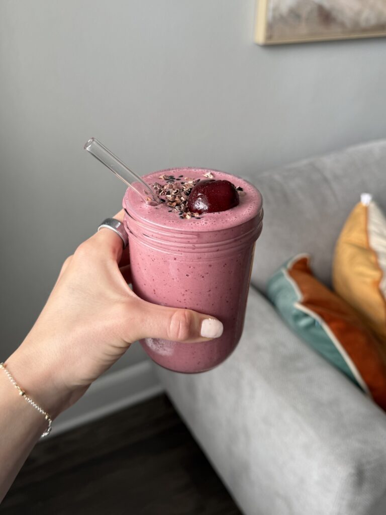 Hand holding a glass jar of cherry tahini smoothie topped with cacao nibs, sesame seeds, and a whole dark cherry, with a glass straw