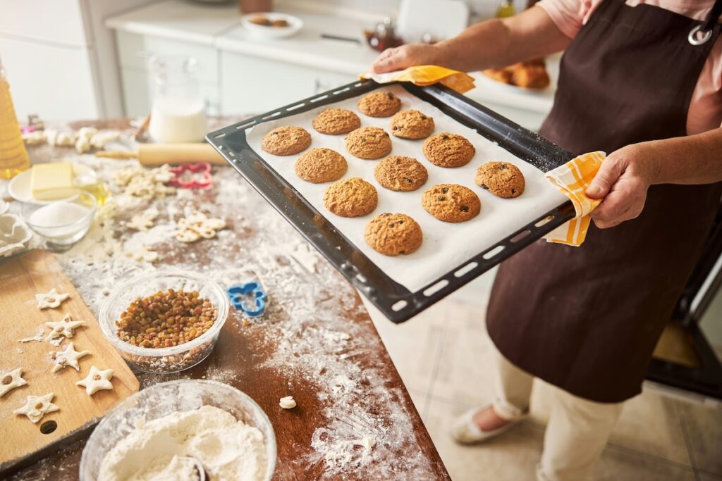 A baker holds a tray of freshly baked oatmeal cookies, one of many healthy fall desserts, over a floured kitchen counter with raisins and baking ingredients.