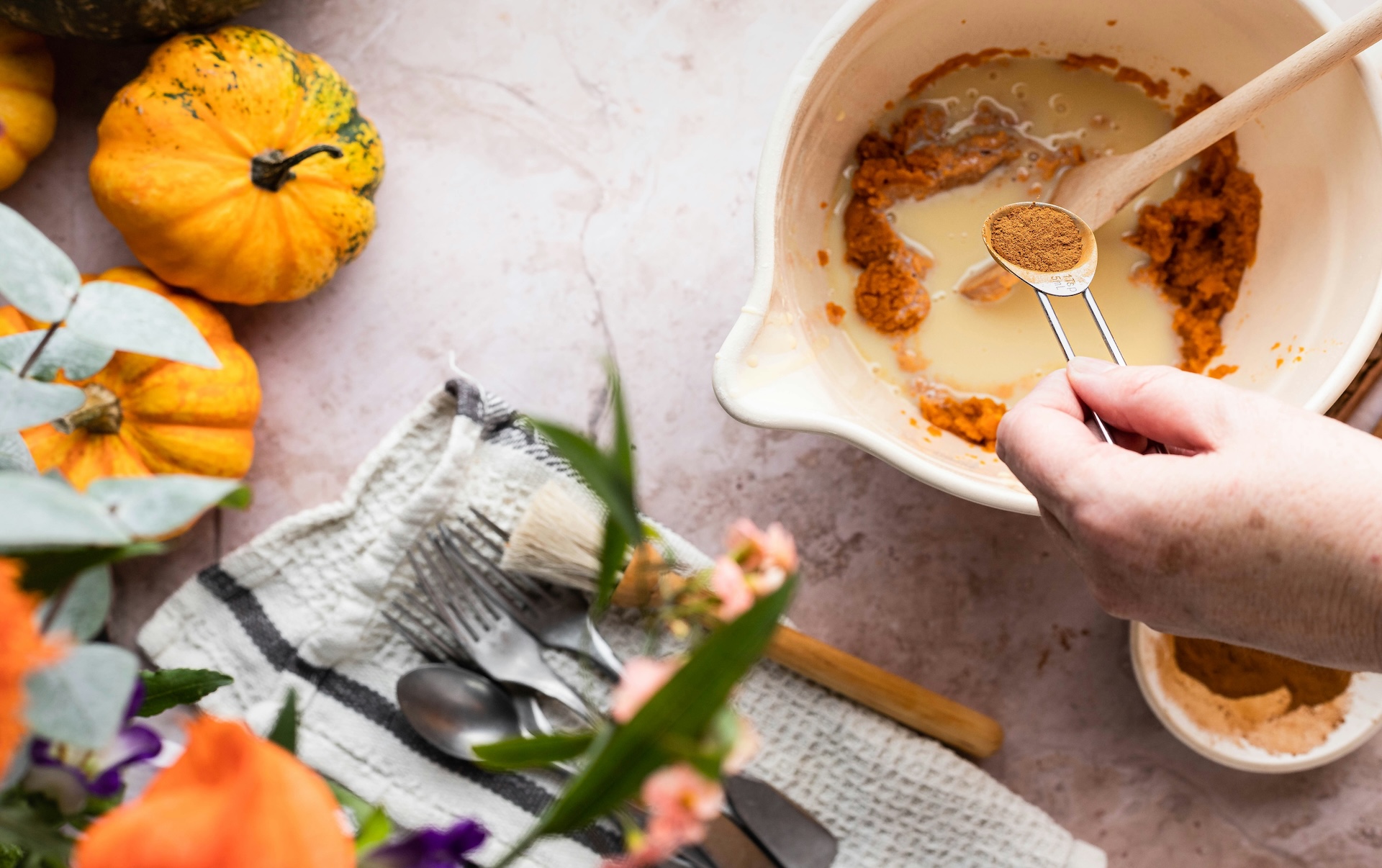 A hand adds a teaspoon of pumpkin spice to a mixing bowl of pumpkin purée and milk, the start of o
