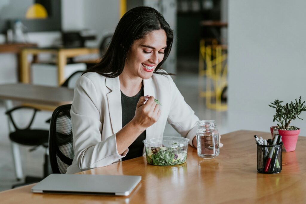 Smiling woman eating a fresh snack at her desk with a glass of water, an example of healthy snacks for work