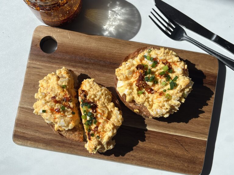 High protein egg salad on toasted bagel halves drizzled with chili oil and topped with green onions on a wooden board