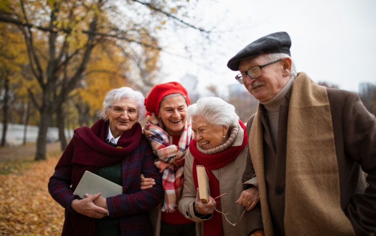 Group of older friends laughing and walking arm in arm through an autumn park, sharing longevity habits like staying social