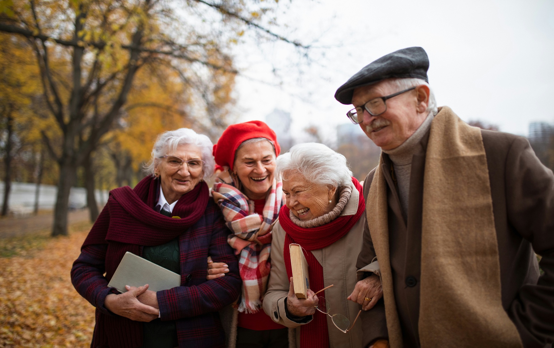 Group of older friends laughing and walking arm in arm through an autumn park, sharing longevity habits like staying social