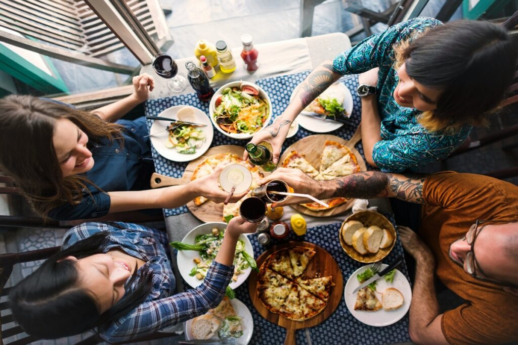 Friends toasting drinks over a shared meal of pizza and salad, a social habit tied to longevity habits