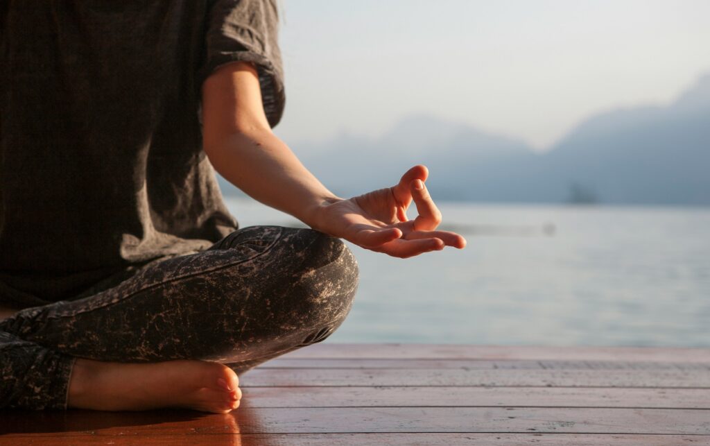 Close-up of a person sitting cross-legged in a meditation pose on a lakeside dock, a daily practice linked to meditation and happiness.