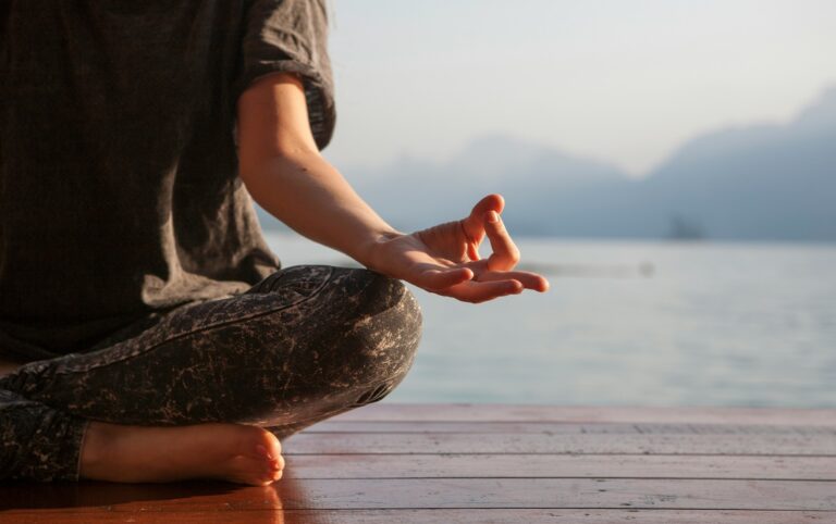 Close-up of a person sitting cross-legged in a meditation pose on a lakeside dock, a daily practice linked to meditation and happiness.