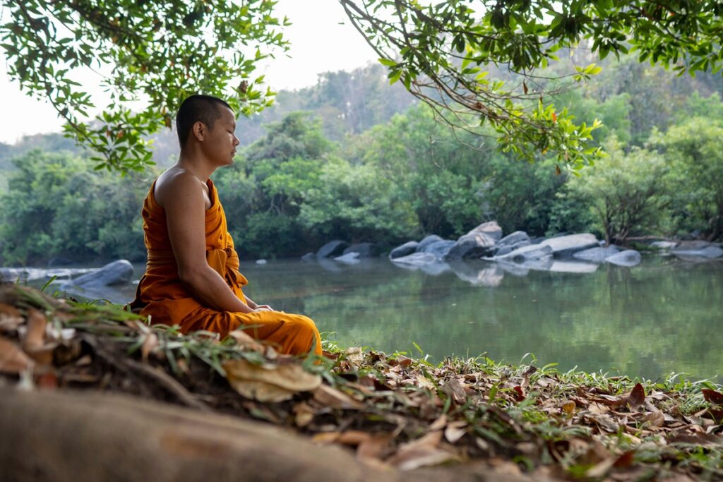 A Buddhist monk meditates beside a calm river, illustrating the link between meditation and happiness.