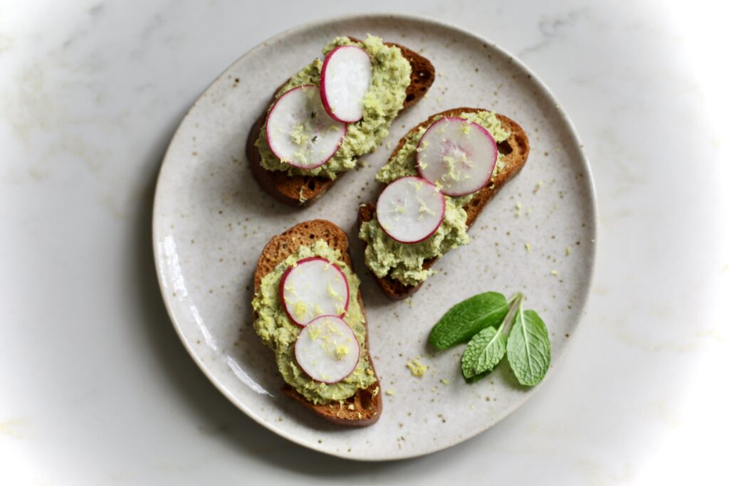 Three crostini topped with pea pesto, sliced radishes, and lemon zest, served with fresh mint leaves