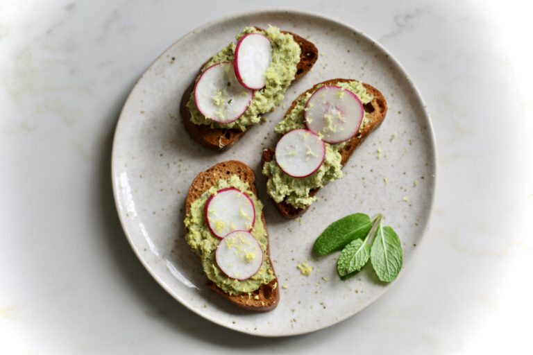 Three crostini topped with pea pesto, sliced radishes, and lemon zest, served with fresh mint leaves