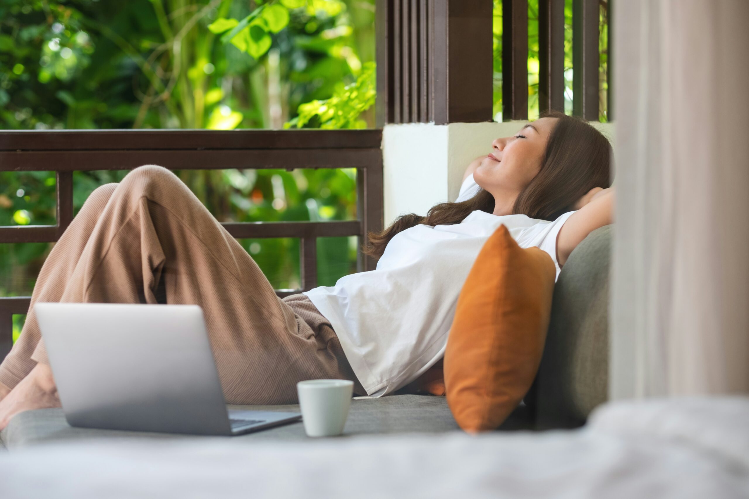 Self-care ideas in action: woman in loungewear relaxing on a porch couch with eyes closed and her laptop set aside