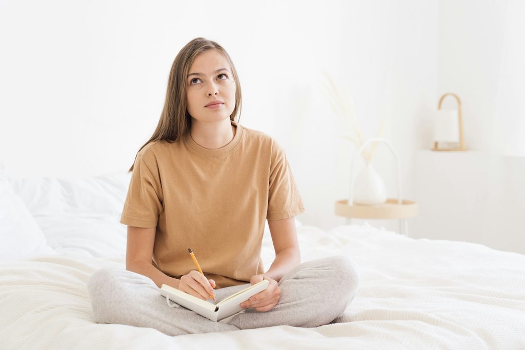 Woman sitting cross-legged on her bed journaling, writing down self-care ideas to build into her daily routine