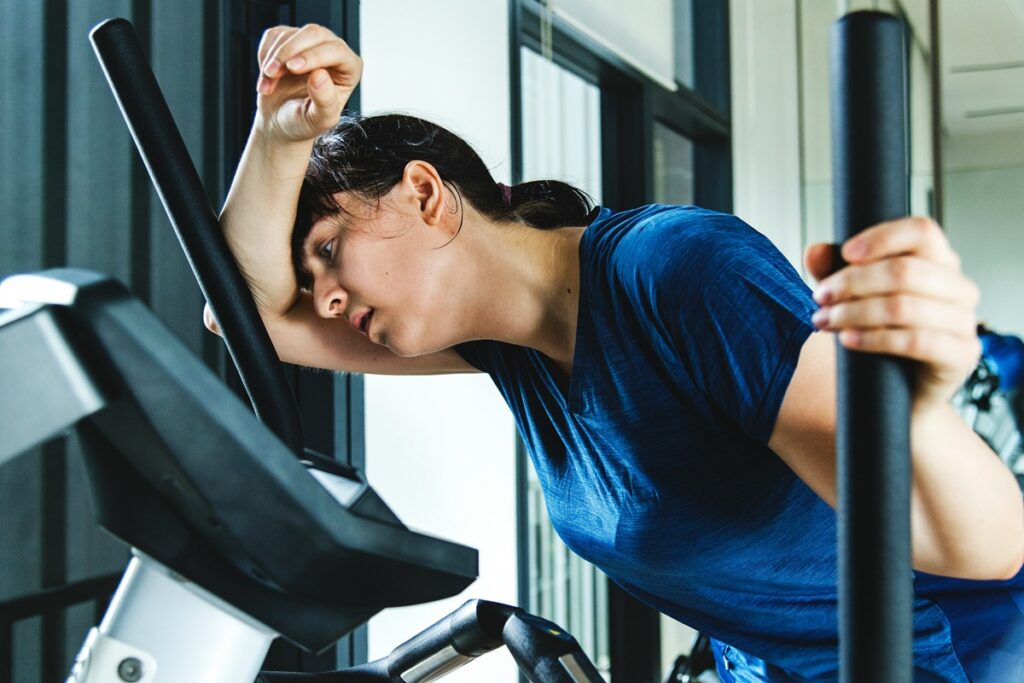 An exhausted woman in a sweaty blue shirt leans her forehead on her arm while on an elliptical machine at the gym, showing the strain of working out while sick.
