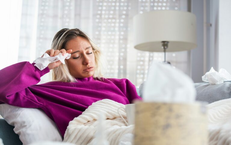 A woman with a cold rests on the couch under a blanket, holding a tissue to her forehead, with a mug and a tissue box nearby. She’s taking a rest day instead of working out while sick.